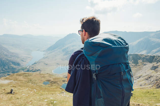 Vista posterior del excursionista masculino con mochila disfrutando del paisaje de las tierras altas durante el trekking en Gales - foto de stock