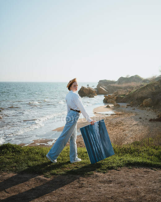 Vista lateral de la joven artista femenina en traje elegante y sombrero caminando cerca de la playa de arena de mar ondulado con pintura en la mano - foto de stock