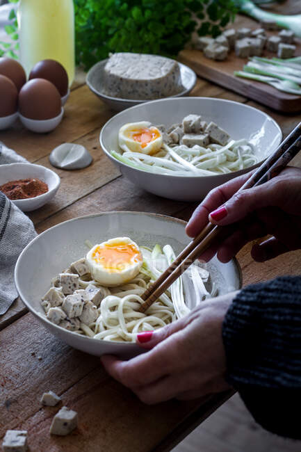Cultivé personne méconnaissable préparant des nouilles ramen fraîches cuites avec du tofu, des œufs et des légumes avec des baguettes sur une table en bois — Photo de stock