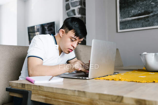 Enfoque adolescente latino con síndrome de Down netbook de navegación mientras está sentado en la mesa y estudiar en línea desde casa - foto de stock