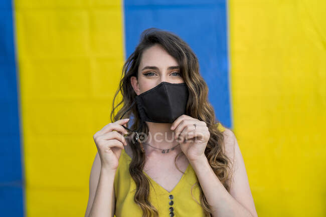 Contenu femelle avec les cheveux ondulés portant un masque de protection pendant le coronavirus en ville en regardant la caméra sur deux fond coloré — Photo de stock