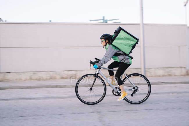 Vista lateral do correio feminino com saco térmico andar de bicicleta na estrada enquanto entrega comida na cidade, borrão de movimento — Fotografia de Stock