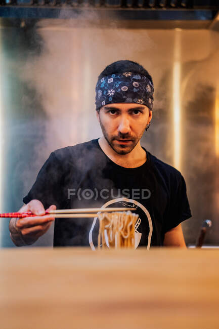 Young bearded male chef in bandana looking at camera while holding noodle with chopsticks in ramen bar — Stock Photo