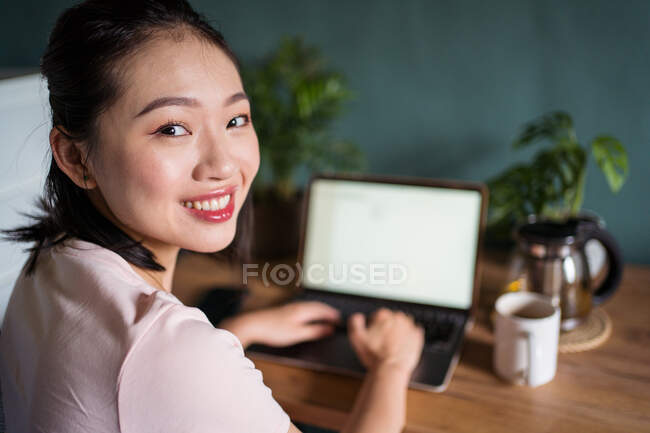 Vue latérale des documents de lecture de pigiste asiatique heureuse sur ordinateur portable tout en étant assis à table pendant le travail à distance en regardant la caméra — Photo de stock