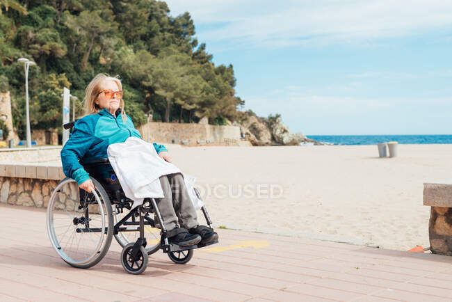 Mujer anciana en silla de ruedas cabalgando a lo largo del terraplén cerca del mar y disfrutando del día de verano - foto de stock