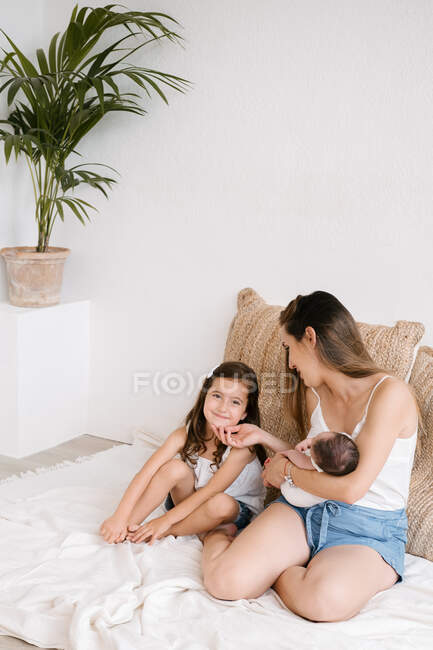 Милая маленькая девочка сидит с нежной матерью, держа новорожденного ребенка, сидя на диване дома — стоковое фото