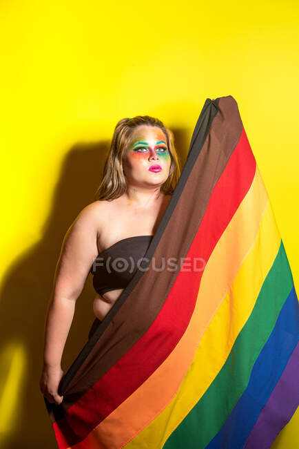 Надлишкова жіноча модель з креативним макіяжем, що показує прапор ЛГБТ і дивиться на жовте тло — стокове фото