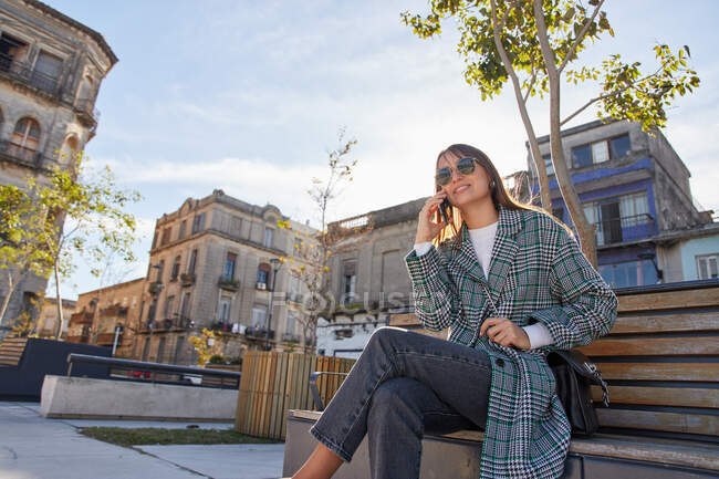 Современная женщина тысячелетия в стильном весеннем наряде сидит на скамейке и отвечает на телефонный звонок во время отдыха на городской улице — стоковое фото