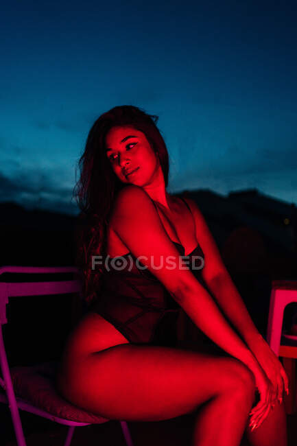 Молода жінка в нижній білизні дивиться далеко, сидячи на стілець під червоним неоновим світлом вночі на терасі — стокове фото