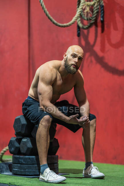 Вид сбоку измученного мускулистого мужчины, смотрящего на камеру, сидящего на гирях и отдыхающего во время функциональной тренировки в тренажерном зале — стоковое фото