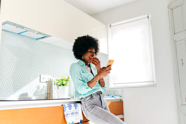 Снизу молодая афроамериканка в повседневной одежде ест яблоко и просматривает интернет по мобильному телефону, стоя на домашней кухне — стоковое фото