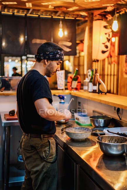 Vista lateral del hombre en bandana de pie en el mostrador y ramen de cocina en la cafetería asiática moderna - foto de stock