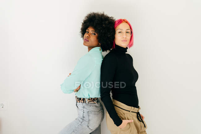 Young pink haired woman and African American girlfriend in stylish outfit standing together back to back on white background — Stock Photo