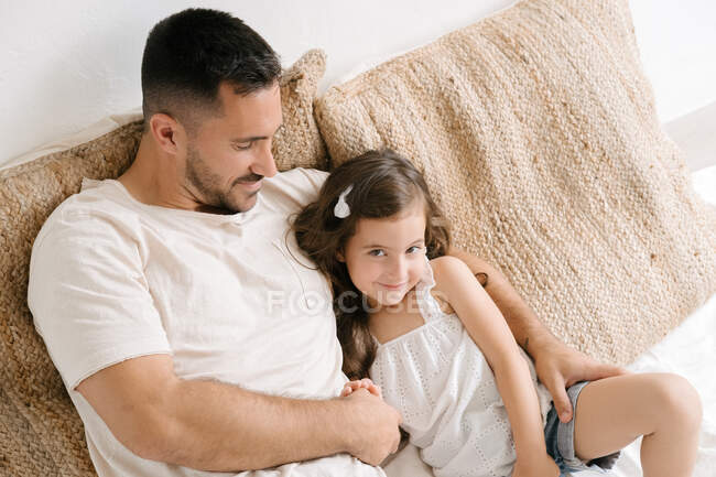 Feliz padre sentado en la cama en casa con adorable niña divirtiéndose en casa mirando hacia otro lado - foto de stock