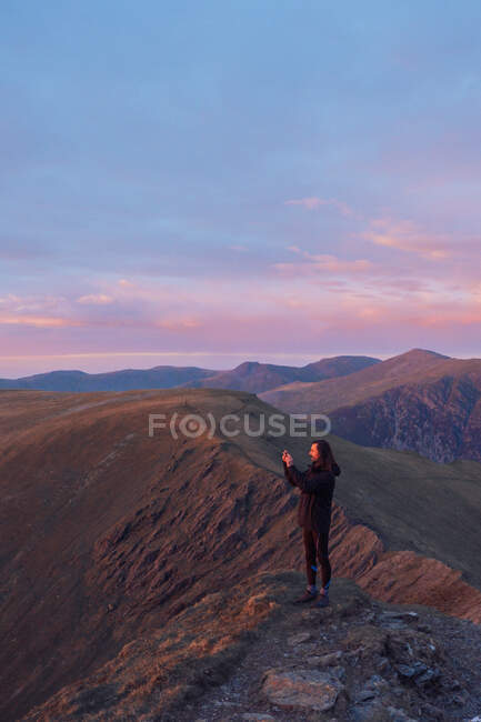 Vista lateral del excursionista masculino tomando fotos de las tierras altas en el teléfono inteligente al atardecer en Gales - foto de stock