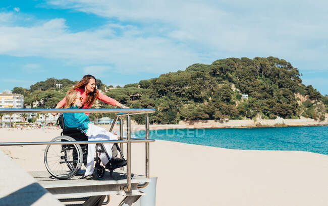 Vista lateral da mulher adulta com mãe idosa em cadeira de rodas admirando vista do mar no dia ensolarado — Fotografia de Stock