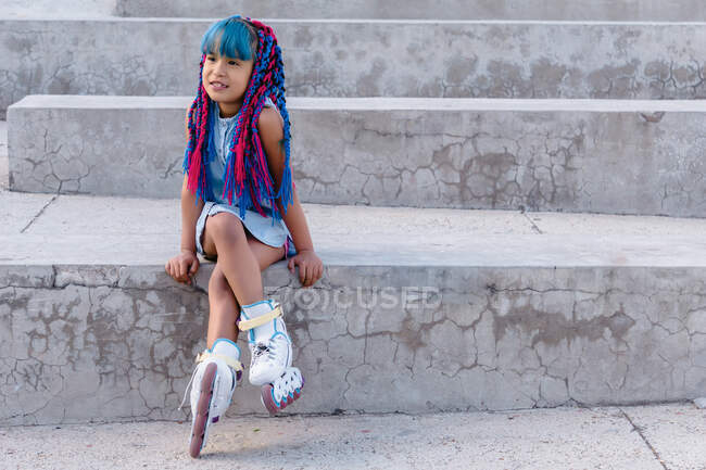 Criança étnica sonhadora com tranças coloridas sentadas com pernas cruzadas na escada enquanto olha para longe à luz do dia — Fotografia de Stock