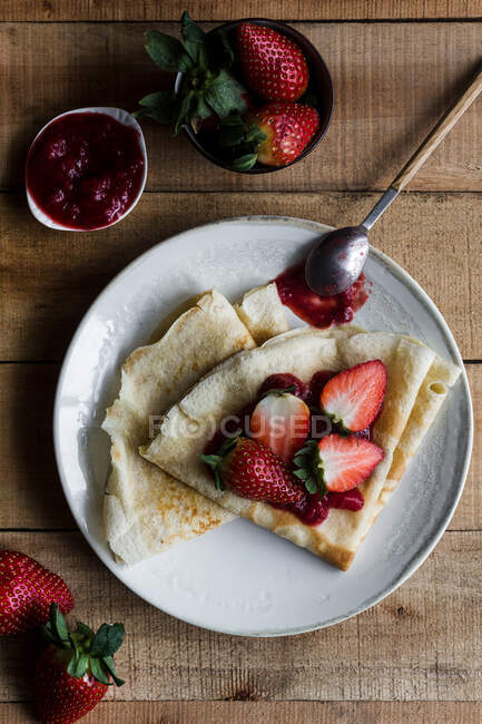 Vista superior de deliciosas crepas con mermelada de fresa dulce colocada en el plato cerca de la cuchara en la mesa de madera - foto de stock