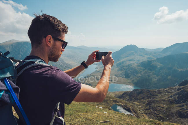 Vue latérale de l'explorateur masculin prenant des photos de montagnes rocheuses sur téléphone portable pendant la randonnée en été au Pays de Galles — Photo de stock