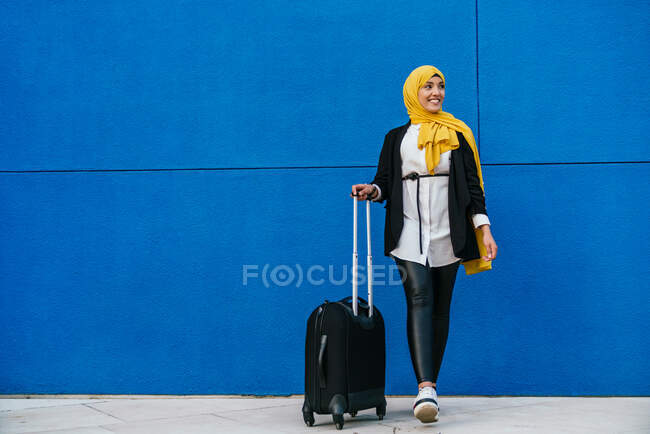 Cheerful Muslim female in headscarf standing with luggage in street against blue wall — Stock Photo
