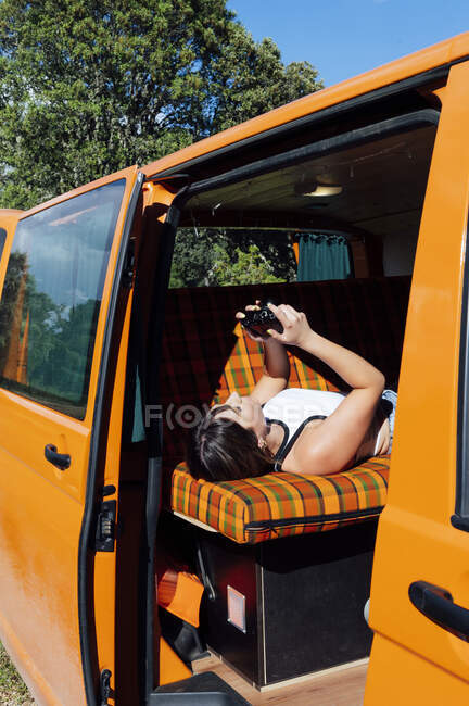 Seitenansicht einer Reisenden, die in einem Lieferwagen liegt und während des Sommerurlaubs durch aufgenommene Bilder der Fotokamera schaut — Stockfoto
