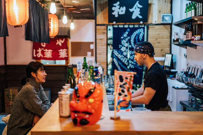 Side view of Asian woman in casual wear sitting at counter and talking with male worker of modern ramen bar — Stock Photo