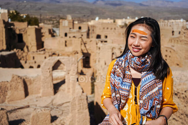 Happy young female tourist in colorful outfit smiling and looking at camera while standing against aged ruined buildings of ancient abandoned Al Hamra village in Oman — Stock Photo
