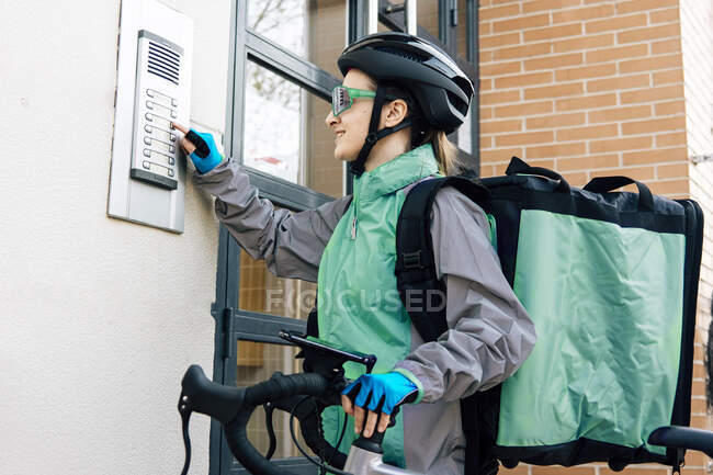 Correio feminino com botão de prensagem de encomendas no dispositivo de intercomunicação na parede de tijolo ao fazer a entrega no dia ensolarado na cidade — Fotografia de Stock