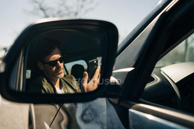 Бородатый мужчина в солнечных очках просматривает мобильный телефон, отражаясь в боковом зеркале автомобиля при солнечном свете — стоковое фото