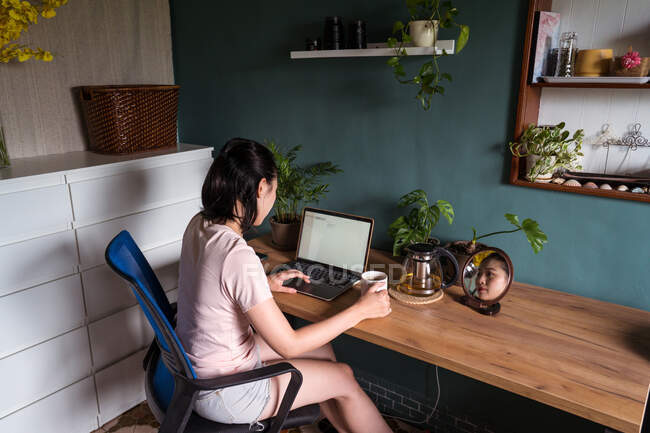 Side view of Asian female freelancer reading documents on laptop while sitting at table during remote work — Stock Photo