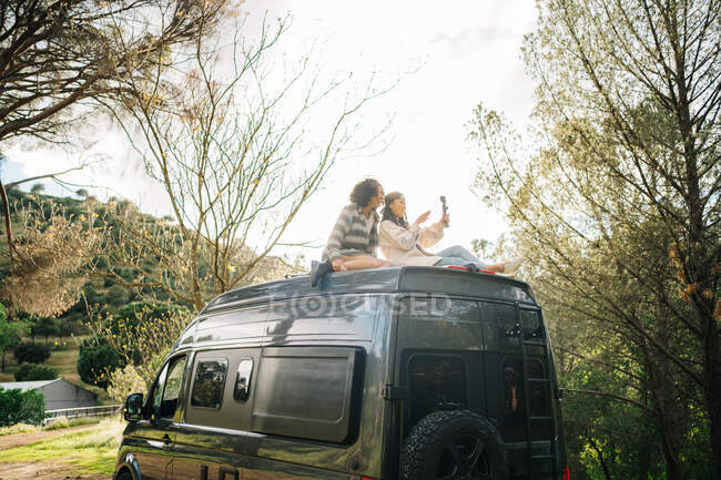 Bajo ángulo de alegres novias jóvenes multirraciales sentadas en el techo de la caravana y tomando fotos mientras pasan las vacaciones de verano juntos en el campo - foto de stock