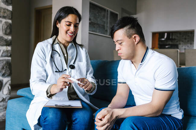 Cheerful female practitioner in medical gown talking to teen boy with Down syndrome and taking notes on clipboard during appointment at home — Stock Photo
