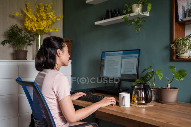 Vista laterale di donne asiatiche freelance che leggono documenti sul monitor del computer mentre siedono a tavola durante il lavoro remoto — Foto stock