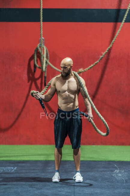 Бородатый парень без рубашки с веревкой на плечах во время интенсивной тренировки в современном спортзале — стоковое фото
