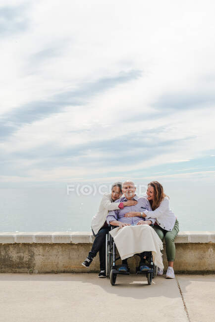 Glückliche Familie umarmt betagten Vater, der an sonnigem Tag im Rollstuhl auf einer Böschung am Meer sitzt — Stockfoto