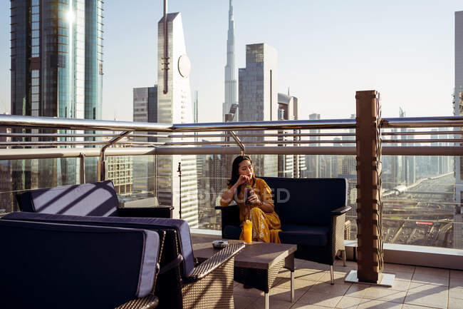 Молодая женщина-путешественница пьет кофе и наслаждается захватывающим видом на Дубай с современной архитектурой, сидя на террасе на крыше современного кафе — стоковое фото