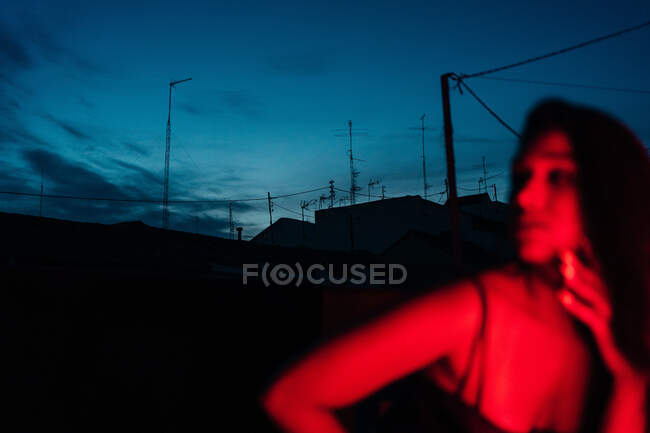 Приємна молода іспанська етнічна жінка в білизні дивиться на камеру, відпочиваючи на терасі під червоним неоновим світлом вночі з синім темним небом. — стокове фото