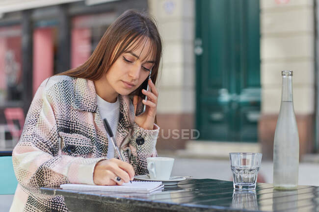 Young woman taking notes in notebook during phone conversation while sitting at table in outdoor cafe in city — Stock Photo