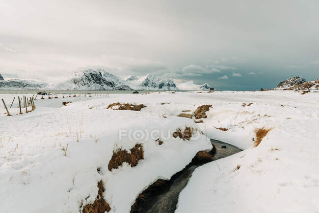 Enger Bach mit kaltem Wasser fließt unter Schnee gegen Berge und bewölkten Himmel auf den Lofoten, Norwegen — Stockfoto
