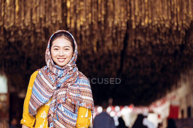 Joven hembra oriental con vestido tradicional y pañuelo en la cabeza sonriendo y mirando a la cámara mientras está de pie sobre un fondo borroso del antiguo bazar zoco de Manama en la ciudad de Manama en Bahréin - foto de stock