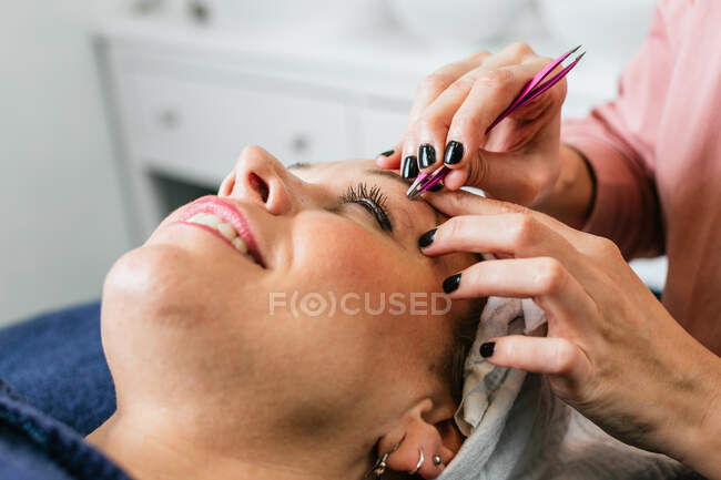 Anonyme Kosmetikerin zupft Augenbrauen im Gesicht einer Kundin, die im Schönheitssalon auf dem Tisch liegt — Stockfoto