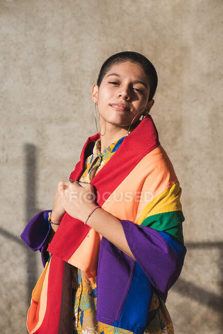 Jovem mulher étnica bissexual com bandeira multicolorida olhando para a câmera e representando símbolos LGBTQ no dia ensolarado — Fotografia de Stock