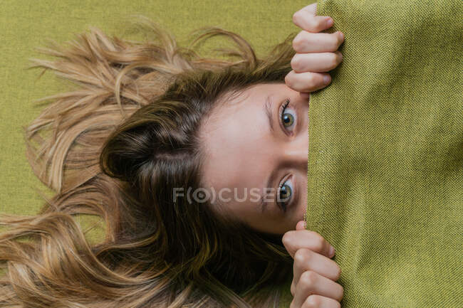 Vue du dessus de la jeune femelle avec de longs cheveux clairs et des yeux verts couvrant la moitié du visage avec un tissu vert et regardant la caméra — Photo de stock