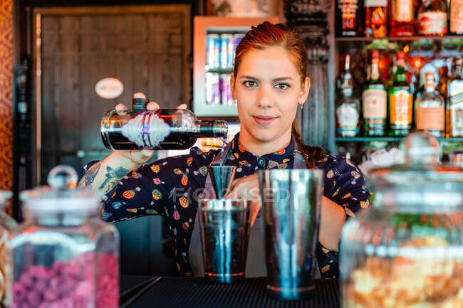Contenuto barista femminile versare bevanda alcolica in metallo shaker mentre si prepara cocktail rinfrescante al bancone nel bar — Foto stock