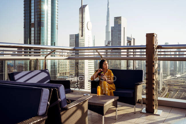 Young female traveler drinking coffee and enjoying breathtaking view of Dubai city with contemporary architecture while sitting on rooftop terrace of modern coffee shop — Stock Photo