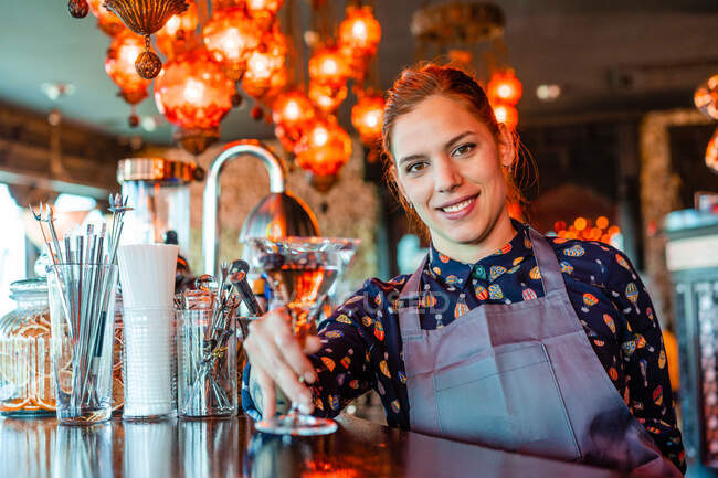Gardienne de bar joyeuse avec un verre de vermouth rafraîchissant debout au comptoir dans le bar et regardant la caméra — Photo de stock