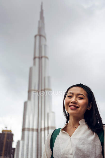 Low angle of smiling young Asian female in casual outfit looking at camera while standing against modern high Burj Khalifa tower in Dubai in cloudy day — Stock Photo