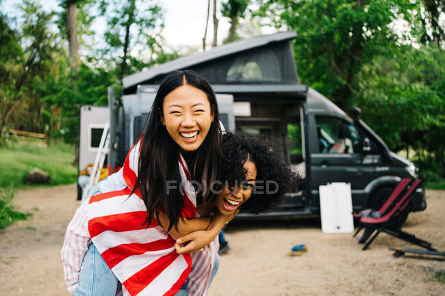 Full length of cheerful young African American woman giving piggyback ride to content Asian girlfriend with American flag while enjoying summer adventure together near camper van in forest — Stock Photo