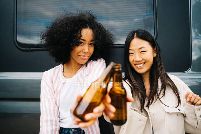 Happy young multiethnic female friends clinking bottles of beer while having fun and enjoying summer holidays together near camper van parked in nature — Stock Photo