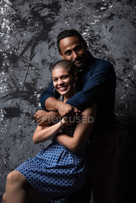 Tender homem étnico abraçando mulher sorridente no fundo escuro no estúdio olhando para a câmera — Fotografia de Stock